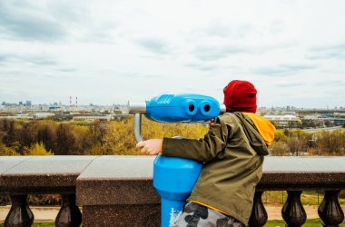 Anaokulu çocuğu şapkalı ve ceketli sokak dürbününün yanında duruyor ve büyük şehre bakıyor. Yerel turizm. Tatildeki çocuklar için eğlence
