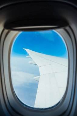 Mavi gökyüzü ve beyaz bulutların arka planında pencere penceresinden geçen uçak kanadı, soğuk renk tonlaması, dikey seyahat içeriği