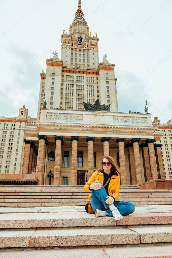 a young female student in a yellow jacket, jeans and glasses sits on the stone steps near the huge university. Getting an education at any age. Age students. Selective Focus