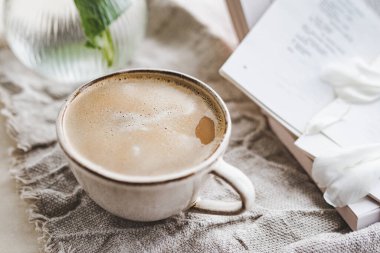 Masada kahve ve köpükle süslü seramik bir fincan, beyaz şakayık ve kitaplarla süslü cam bir vazo. İçkinin atmosferik görüntüsü. seçici odak