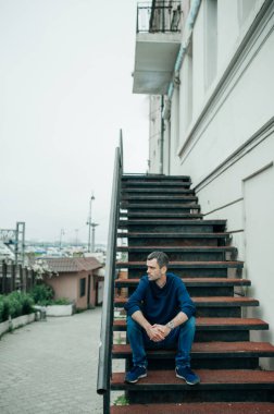 Mavi elbiseli, orta yaşlı, yakışıklı genç bir adam şehrin ortasında merdivenlerde oturuyor. seçici odak