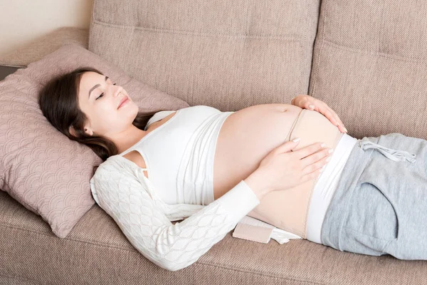 腹部支持包帯にソファの上に寝そべっている妊婦 マタニティヘルスケアのコンセプトサポート製品 — ストック写真