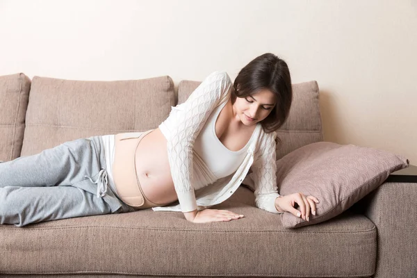 腹部支持包帯にソファの上に寝そべっている妊婦 マタニティヘルスケアのコンセプトサポート製品 — ストック写真