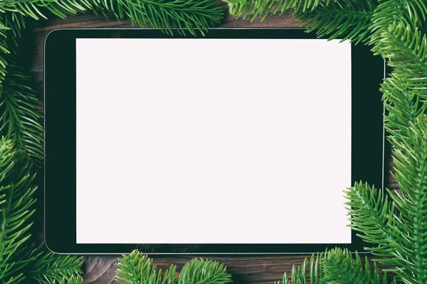 Top view of tablet decorated with a frame made of fir tree on wooden background. New Year time concept.