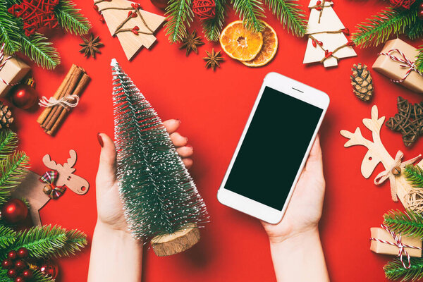 Top view of female hands holding phone in one hand and christmas tree in another hand on red background. New Year holiday concept. Mockup.