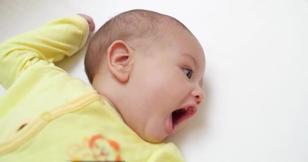Ένα ωραίο μικρό καυκάσιο νεογέννητο μωρό είναι αστείο χαμογελαστό, ξαπλωμένο στο κρεβάτι του παιδιού. Όμορφο χαμογελαστό μωρό στο κρεβάτι στο σπίτι — Αρχείο Βίντεο