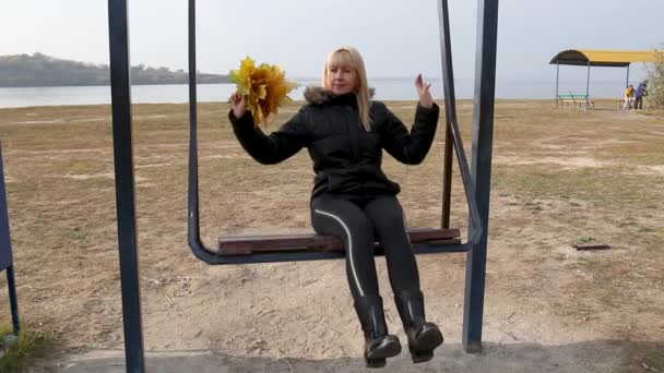 Kobieta relaksuje się na huśtawce w jesienny dzień. kobieta z bukietem żółtych liści jest jazda huśtawka — Wideo stockowe
