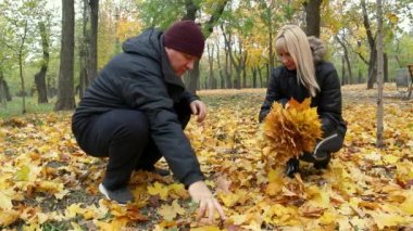 Neşeli adam ve kadın, sonbahar parkında kuru sarı yaprakların üzerinde oturuyorlar ve sarı sonbahar yapraklarını topluyorlar.