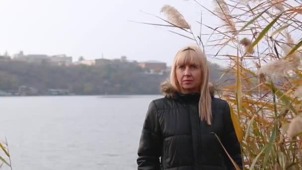 Een vrouw staat op de oever van de rivier in het riet en denkt na. herfststemming en depressie — Stockvideo