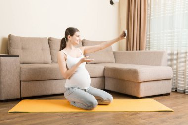 Evde yoga minderinde otururken elinde akıllı telefon tutan hamile bir kadın. Coronavirus zamanında spor ya da yoga eğitimi çevrimiçi.