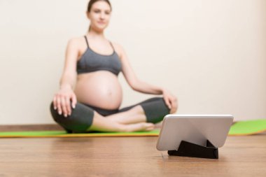Evde yoga minderinde otururken elinde akıllı telefon tutan hamile bir kadın. Coronavirus zamanında spor ya da yoga eğitimi çevrimiçi.
