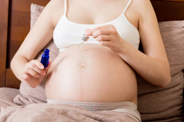 a pregnant girl sits at home on the bed and smears smile an anti-stretch mark cream on her stomach. Pregnancy, motherhood, preparation and expectation concept.