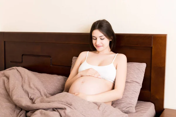 妊娠中の女性はベッドに座って家で腹を触っていました 妊娠期待の概念 — ストック写真