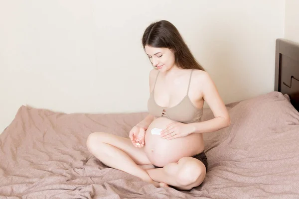 一个怀孕的女孩坐在家里的床上 在她的肚子上涂了一个抗拉伸标记霜 准备和期望概念 — 图库照片