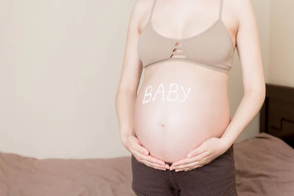 一个怀孕的女孩坐在家里的床上 在她的肚子上涂了一个抗拉伸标记霜 准备和期望的概念 肚子上的婴儿一词 — 图库照片