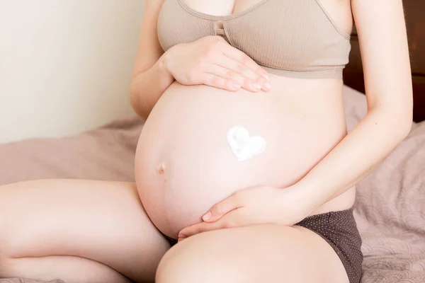 一个怀孕的女孩坐在家里的床上 在她的胃部涂上了抗拉伸标记霜 准备和期望概念 — 图库照片