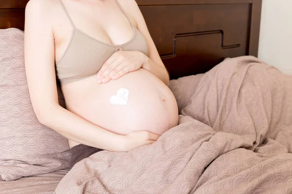 一个怀孕的女孩坐在家里的床上 在她的胃部涂上了抗拉伸标记霜 准备和期望概念 — 图库照片