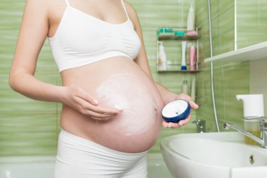 Genç hamile bir kadın sabah banyoda karnına gerilme önleyici krem sürüyor. Hamilelik, annelik ve vücut bakımı.