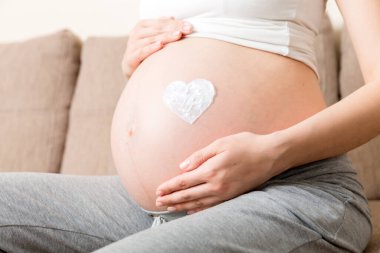 Hamile bir kız evde yatağında oturuyor ve kalbine esneme önleyici kremi sürüyor. Hamilelik, annelik, hazırlık ve beklenti konsepti.