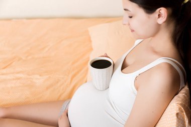 Hamile genç bir kadın yatakta oturuyor ve evde kahve içiyor. annelik kavramı.