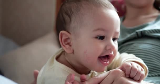 一位年轻的母亲抱着一个漂亮的7个月大的儿子，她笑着咬着手指和母亲的手。新生儿的发痒的牙齿，牙疼 — 图库视频影像