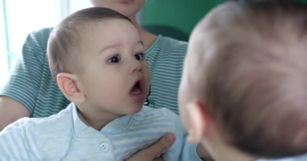 Un lindo bebé recién nacido de siete meses juega con un espejo en casa con su madre en sus brazos, ve su reflejo y abofetea sus palmas en el espejo — Vídeos de Stock