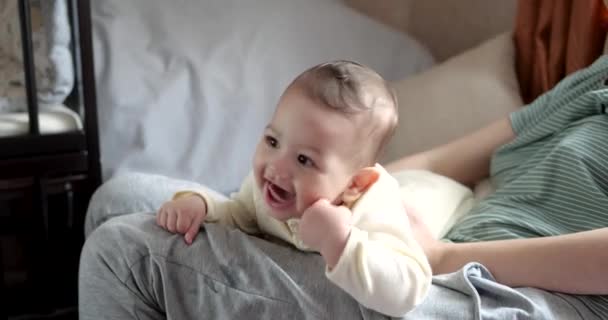 Een jonge moeder houdt een mooie zeven maanden oude zoon vast die glimlacht en knaagt aan vingers en moeders hand. jeukende tanden van een pasgeborene, tandjes krijgen — Stockvideo
