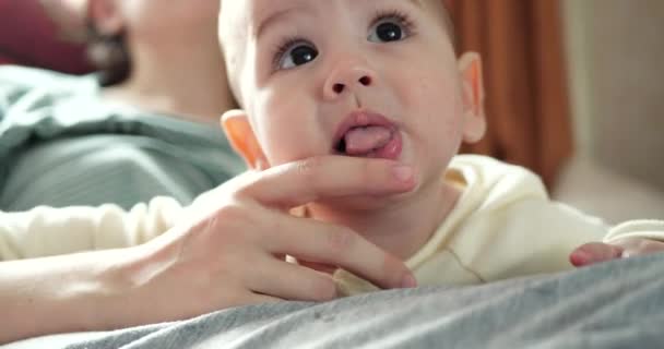 Uma jovem mãe está segurando um belo filho de sete meses de idade que sorri e rói os dedos e as mães mão. comichão nos dentes de um recém-nascido, dentição — Vídeo de Stock