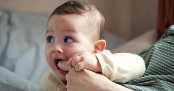 Eine junge Mutter hält einen schönen sieben Monate alten Sohn, der lächelt und an Fingern und Müttern nagt. juckende Zähne eines Neugeborenen, Kinderkrankheiten — Stockvideo