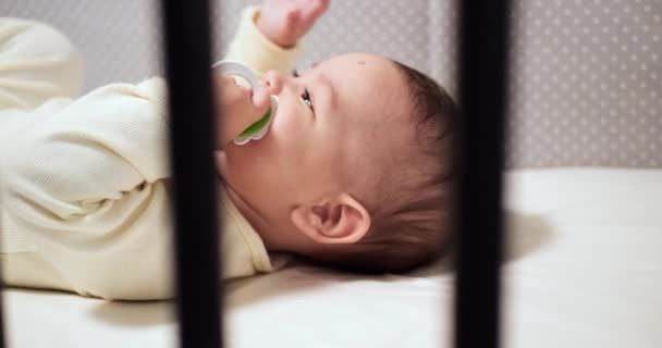 Pasgeboren vrolijke kleine baby kauwt en speelt in zijn wieg met tepel. Grappig glimlachen kijkt door de reling van de wieg. slow motion — Stockvideo