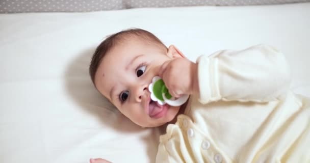 Bayi kecil yang baru lahir yang ceria mengunyah dan bermain di tempat tidurnya dengan puting susu. Lucu tersenyum terlihat melalui pagar tempat tidur bayi. gerak lambat — Stok Video