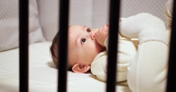 Petit bébé joyeux nouveau-né mâche et joue dans sa crèche avec le mamelon. Drôle de sourire regarde à travers la balustrade de la crèche. au ralenti — Video