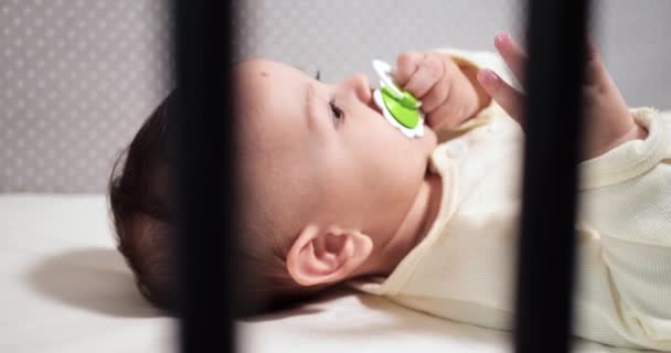 Новорожденный веселый малыш жует и играет в своей кроватке с соском. Смешная улыбка смотрит сквозь перила кроватки. замедленное движение — стоковое видео