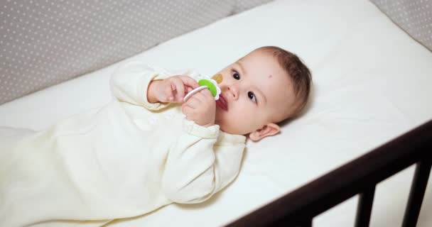 Newborn cheerful littlebaby is chewing and playing in his crib with nipple. Funny smiling looks through the railing of the crib. slow motion — Stock Video