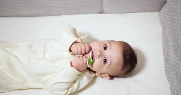 Novorozené veselé děťátko žvýká a hraje si v postýlce s bradavkou. Legrační úsměv se dívá přes zábradlí postýlky. zpomalený pohyb — Stock video