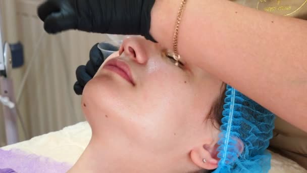 Cosmetólogo está aplicando mascarilla peeling en la cara de las mujeres usando cepillo en clínica de belleza. Esteticista haciendo el procedimiento de cuidado de la piel. Tratamiento de spa en cosmetología — Vídeos de Stock