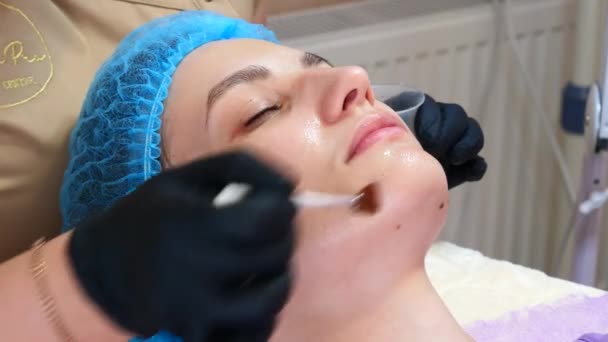 美容クリニックでは、女性の顔にブラシを使って剥離マスクを適用しています。美容師はスキンケア手順を作る。美容室でのスパトリートメント — ストック動画