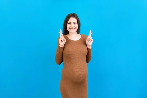 Cheerful young pregnant woman holding fingers crossed, waiting pregnancy isolated on colored background in studio. lifestyle pregnancy concept.