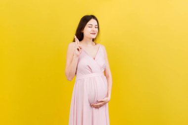 Neşeli genç hamile bir kadın parmaklarını çaprazlayarak stüdyoda izole bir şekilde hamileliğini bekliyor. Yaşam tarzı hamilelik kavramı.