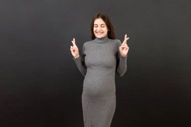 Neşeli genç hamile bir kadın parmaklarını çaprazlayarak stüdyoda izole bir şekilde hamileliğini bekliyor. Yaşam tarzı hamilelik kavramı.