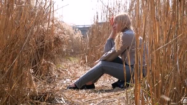 Een vrouw zit bedachtzaam langs het rietveld bij de rivier, heldere dag, blauwe lucht — Stockvideo