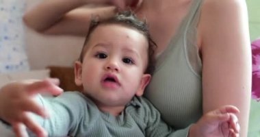 Annesinin kollarında oturmuş gülümseyen bir çocuğun portresi. Parmağını kameraya dayar ve elinden almaya çalışır.