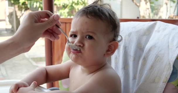 Portret van een kind dat babyvoeding eet met zijn lepel. moeder voedt het kind buiten aan de kindertafel — Stockvideo