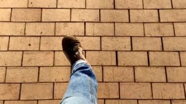 Kahverengi bir kaldırım boyunca yürüyen bir adamın bacaklarının üstünden bak. Spor ayakkabılı ve kot pantolonlu bir adam kaldırımda yürüyor, tepeden manzaralı..