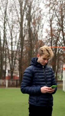 Çevrimiçi haber okuyan bir adam olarak dikey video ve spor sahasındaki akıllı telefondan sosyal ağda SMS yazıyor. Mavi ceketli genç adam sonbahar spor sahasında cep telefonu kullanıyor..