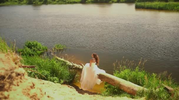 Une mariée romantique en robe de mariée blanche touchant ses longs cheveux bruns près de la rivière — Video