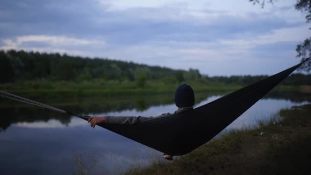 Un hombre cansado en un sombrero mirando el río en una hamaca — Vídeos de Stock