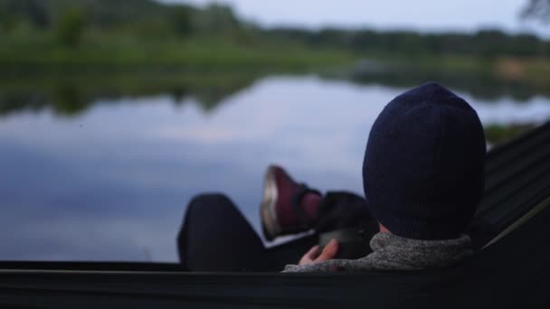 Um viajante experiente em um chapéu comendo nozes na margem do rio — Vídeo de Stock