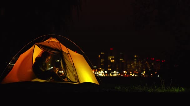 Młody facet w kurtce siedzi w pomarańczowym namiocie i patrzy na smartfona — Wideo stockowe