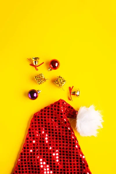 Santa Claus klobouk s vánoční koule, dárky a jiskřivé zvony na žlutém pozadí. — Stock fotografie